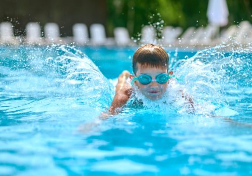 Boy in goggles swimming splashing in pool Pleasure, swimming. Boy in swimming goggles vigorously swimming splashing in pool outdoors on fine day